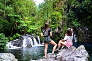 Two girls sit on rocks beside a cascading waterfall, discover a hidden waterfall, and enjoy the natural beauty around them.