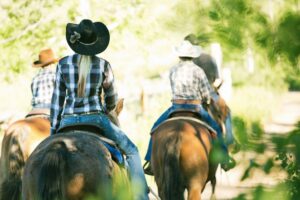 Three people wearing cowboy hats on Horseback riding in Makawao, trotting down a dirt path lined with trees.