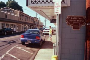 A blue car is stationary on the side of the road in Lahaina Historic District, with a backdrop of trees and a bright sky.