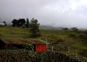 A vibrant red barn is perched on a grassy hill in upcountry Maui, contrasting with the blue sky and distant landscape.